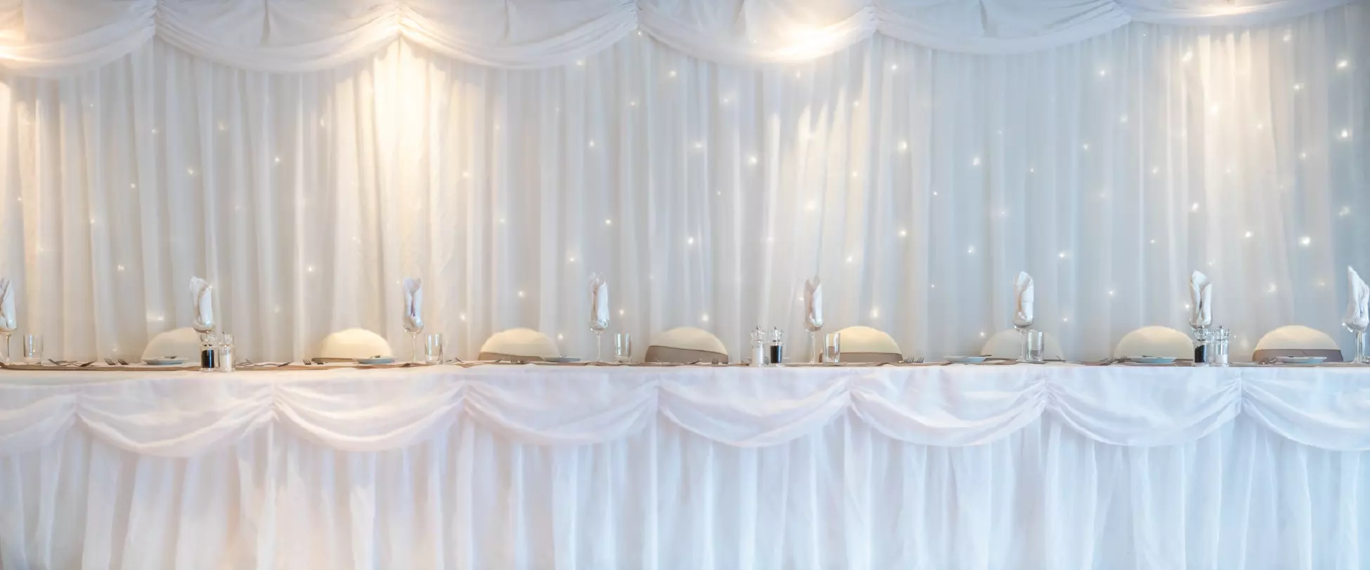 A table set up for the bridal party at a wedding breakfast. The table is covered with white table, white ribbons, and there is a white draping cloth behind the chairs with lights..