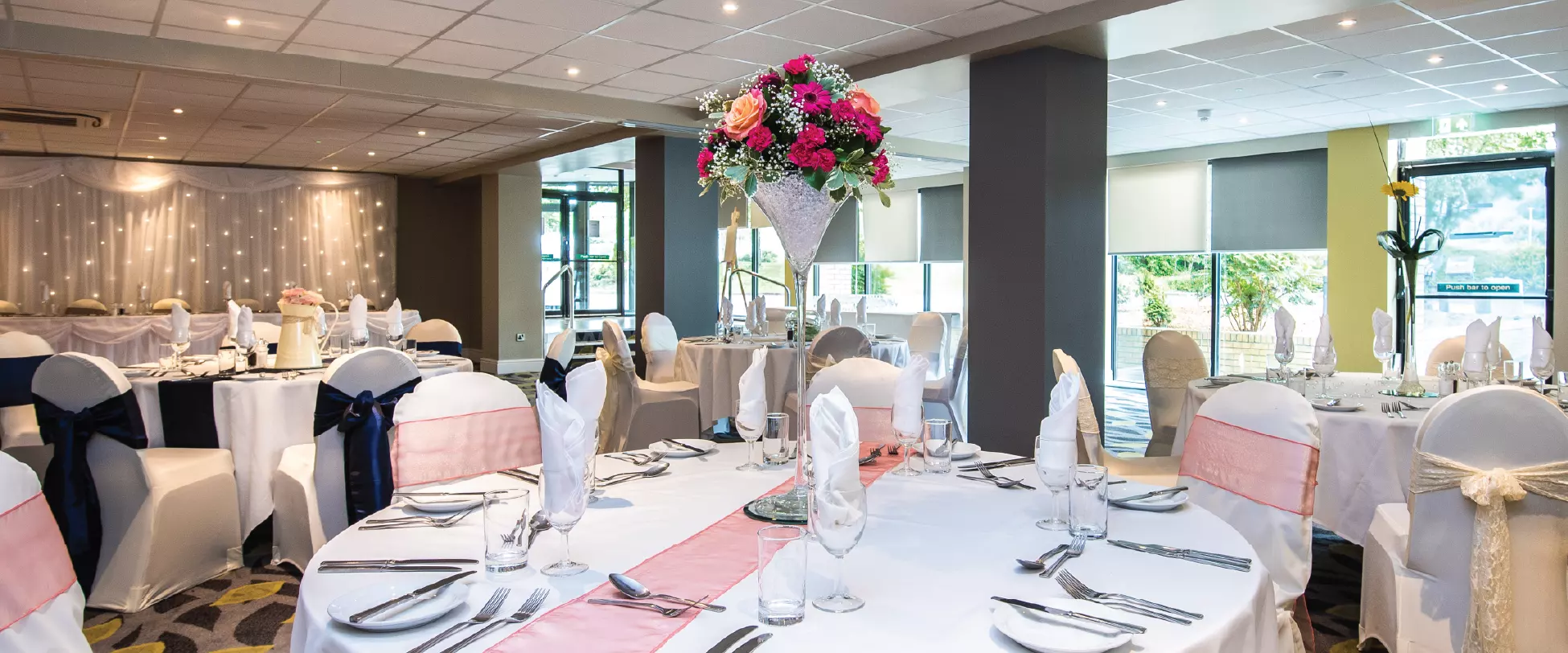 A room set up for a wedding breakfast. Tables are set with white table cloths, pink ribbons and floral centrepieces.