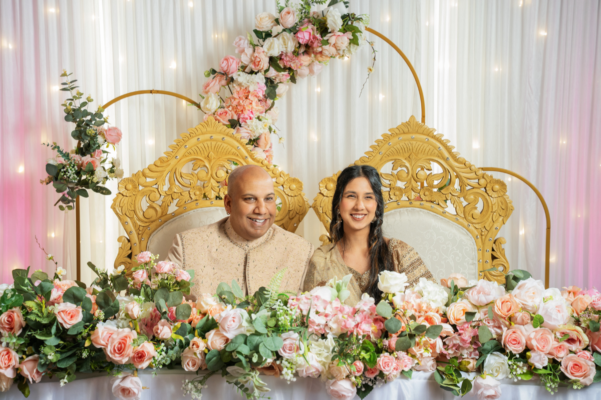 A couple sit in large gold and cream chairs behind a table laid with white linens and a huge garland of pink and white flowers. Behind them is a lit backdrop and even more pink and white flowers..